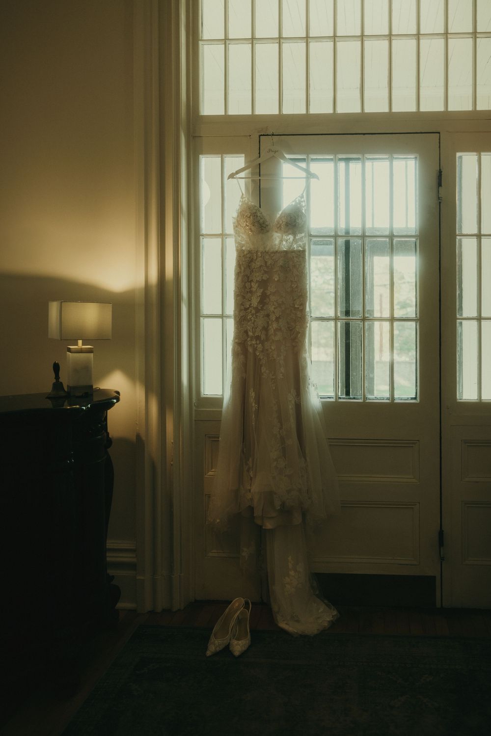 A wedding dress hangs elegantly by a sunlit window creating a dreamy silhouette.