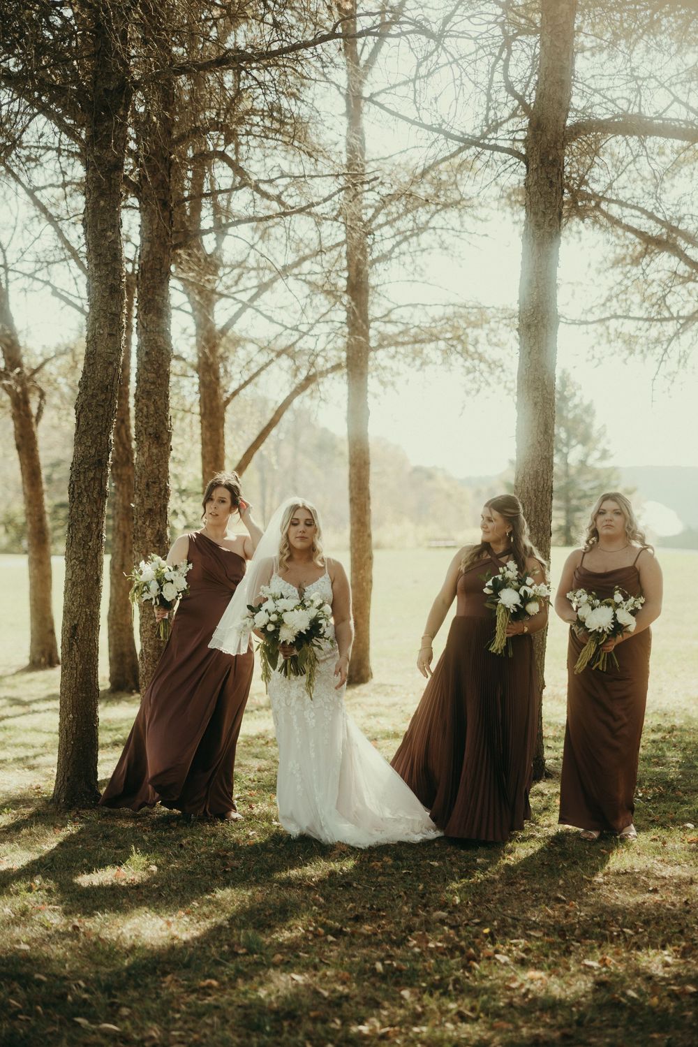 Wedding party poses among tall trees with bridesmaids in brown dresses holding white bouquets.