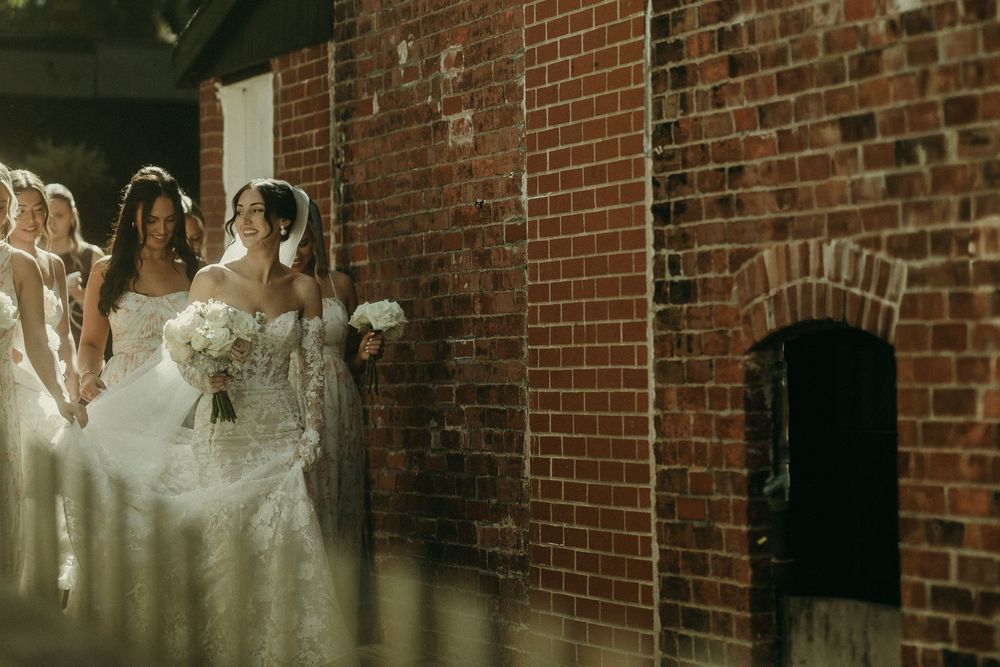 A bride in a white lace gown stands against a rustic brick wall background.