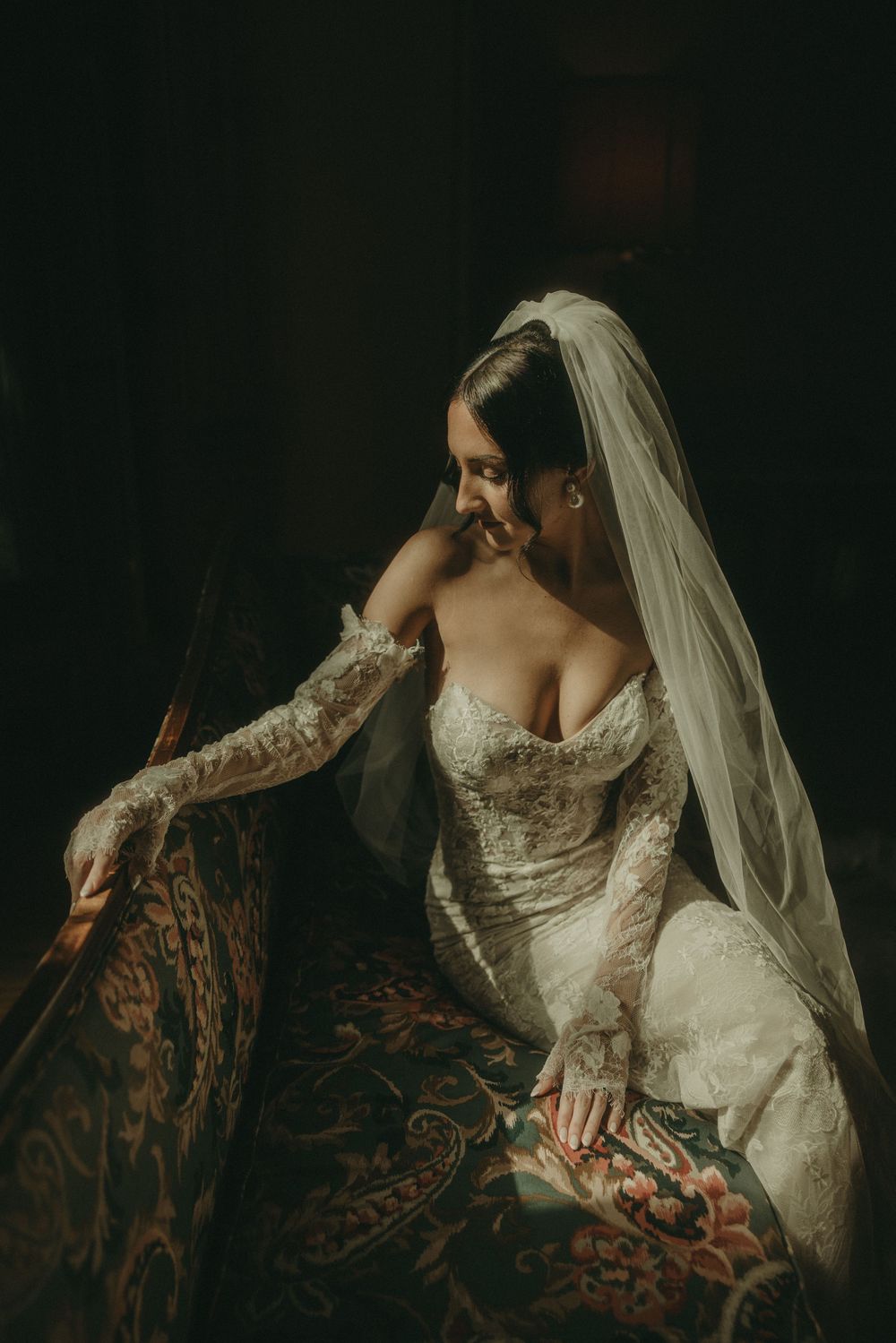 Detailed lace wedding gown and veil captured in moody, dramatic lighting on antique furniture.