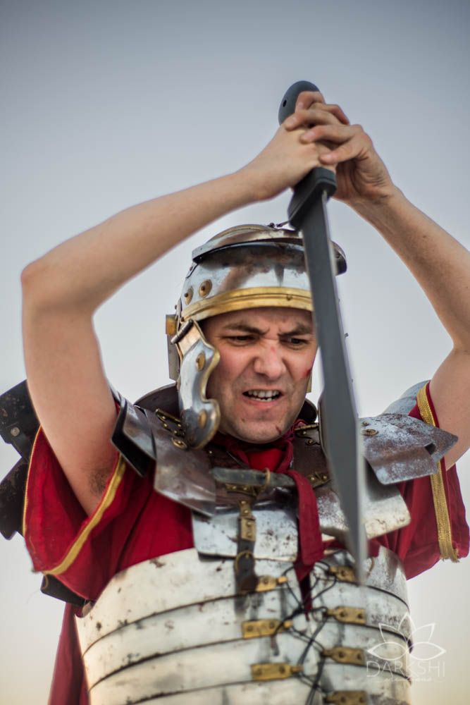 A Roman soldier in full armor raises a sword overhead, grimacing intensely during a historical reenactment.