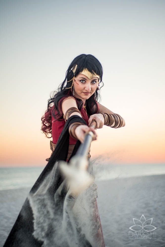 Woman cosplaying as Wonder Woman poses on a beach at sunset, extending her lasso toward the camera.