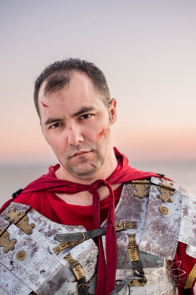 Man in Roman soldier costume with red cape and armor, wearing battle makeup, posing outdoors at sunset.