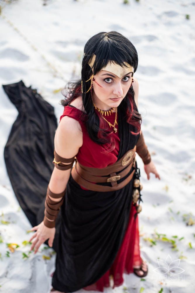 Woman in red and black fantasy cosplay costume with gold jewelry poses dramatically on white sand beach.