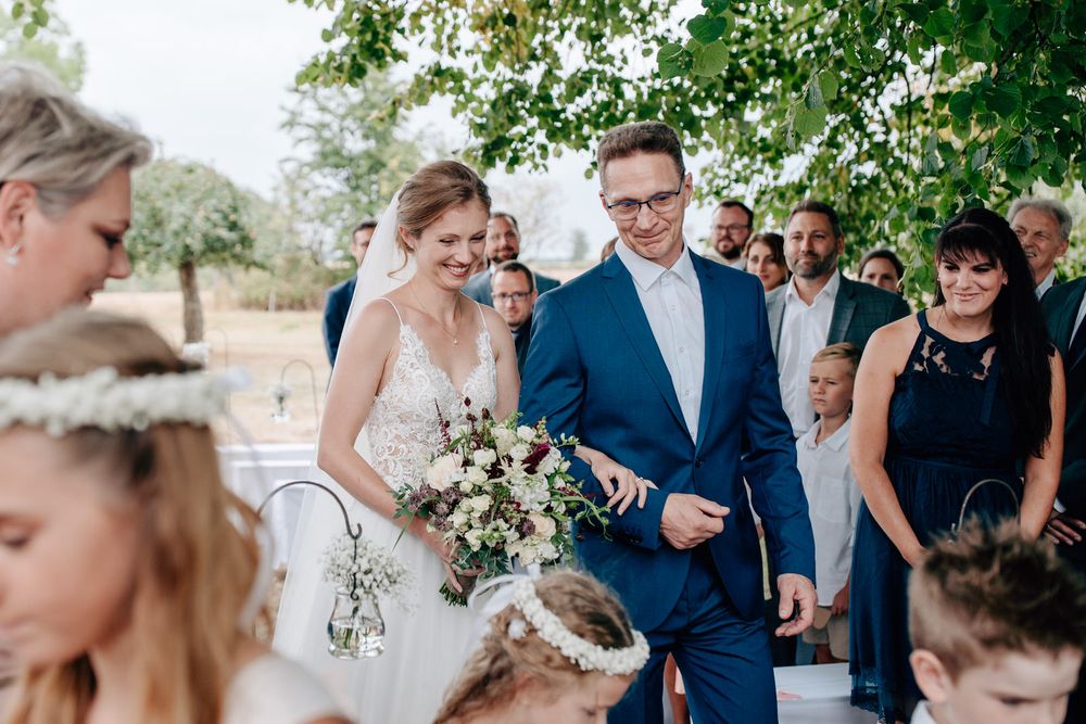 A couple walks down a sunlit path after their wedding ceremony, surrounded by guests in a natural outdoor setting.Weiherhof Wächtersbach