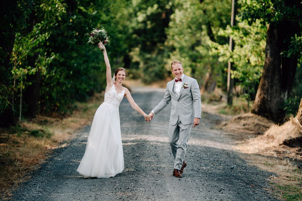 A couple in wedding attire walk and laugh together on a tree-lined gravel path in natural outdoor setting.