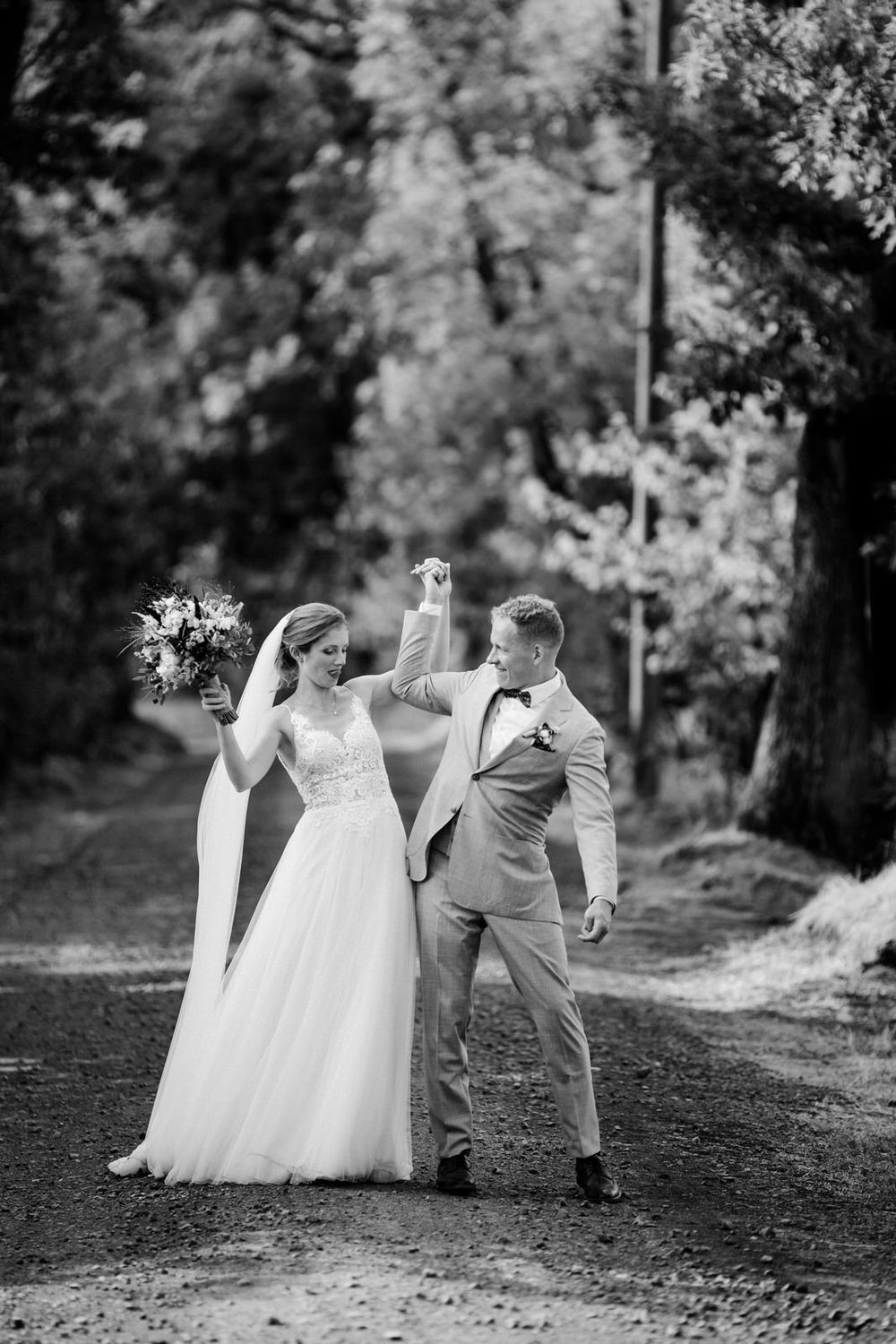 A black and white wedding photo showing a couple celebrating outdoors on a wooded path with raised hands and flowers.