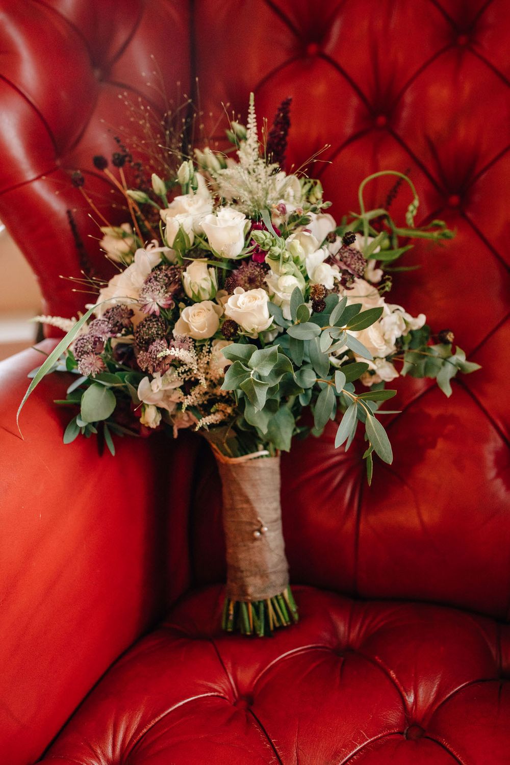 Beautiful rustic wedding bouquet with white roses and eucalyptus resting on red tufted leather seating.Weiherhof Wächtersbach