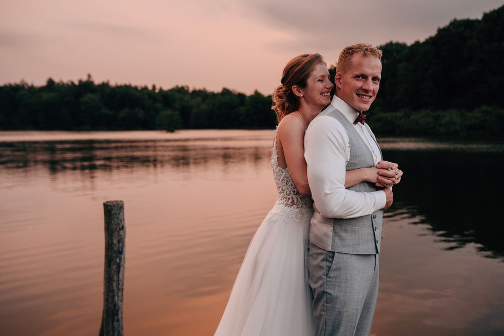 Newlywed couple embraces on wooden dock by serene lake at sunset with pink sky reflecting on calm water.Weiherhof Wächtersbach