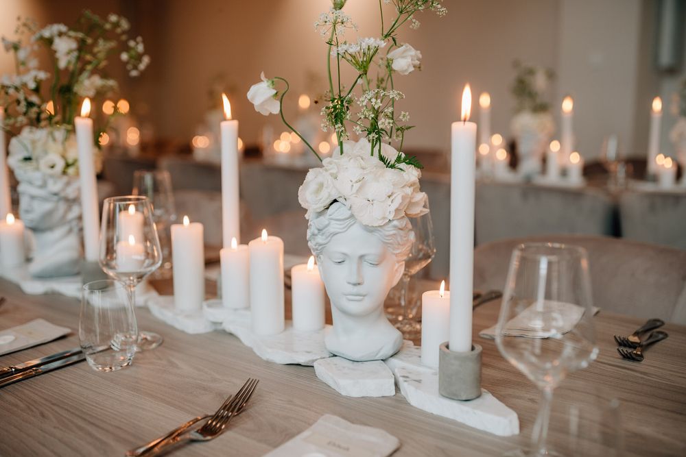 Elegant table setting with white classical bust planter, tall white candles and delicate flowers on rustic wooden surfac la vette ortenberg