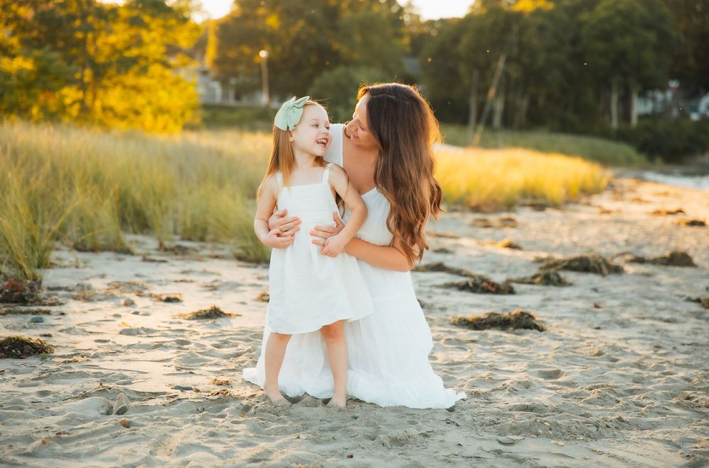 A mother in a white dress kisses her laughing little girl on a sandy beach at golden hour in Portsmouth, RI.