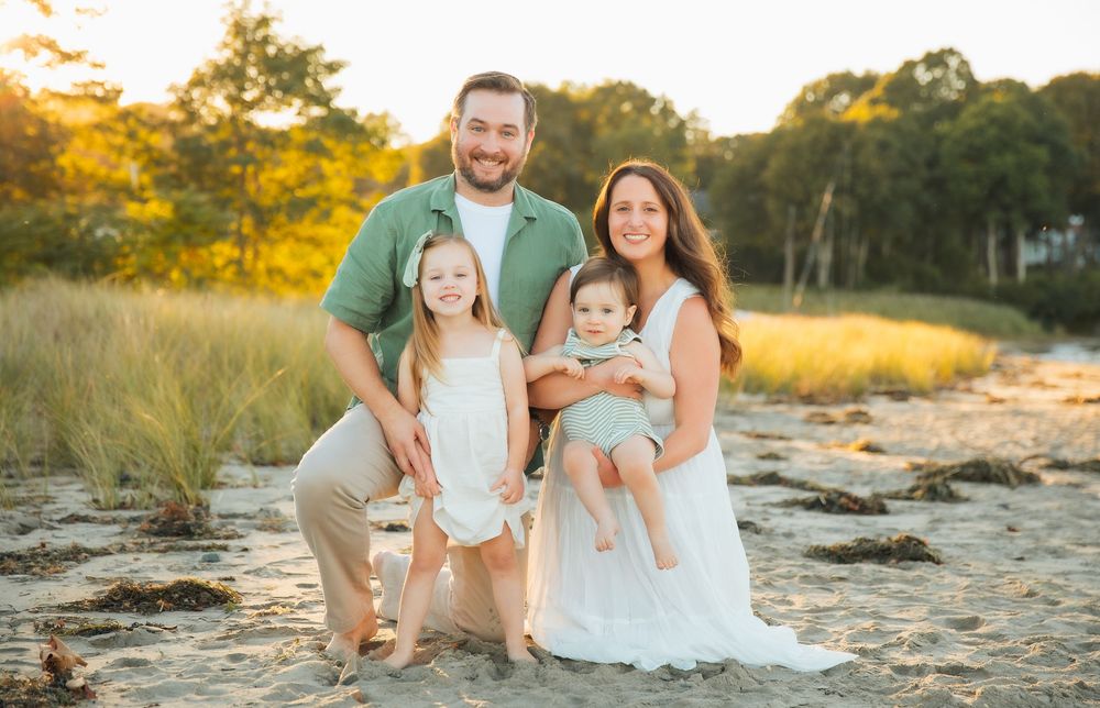 Family of four posing on sandy beach at golden hour, parents with two young children smiling outdoors. Portsmouth, RI Rhode Island
