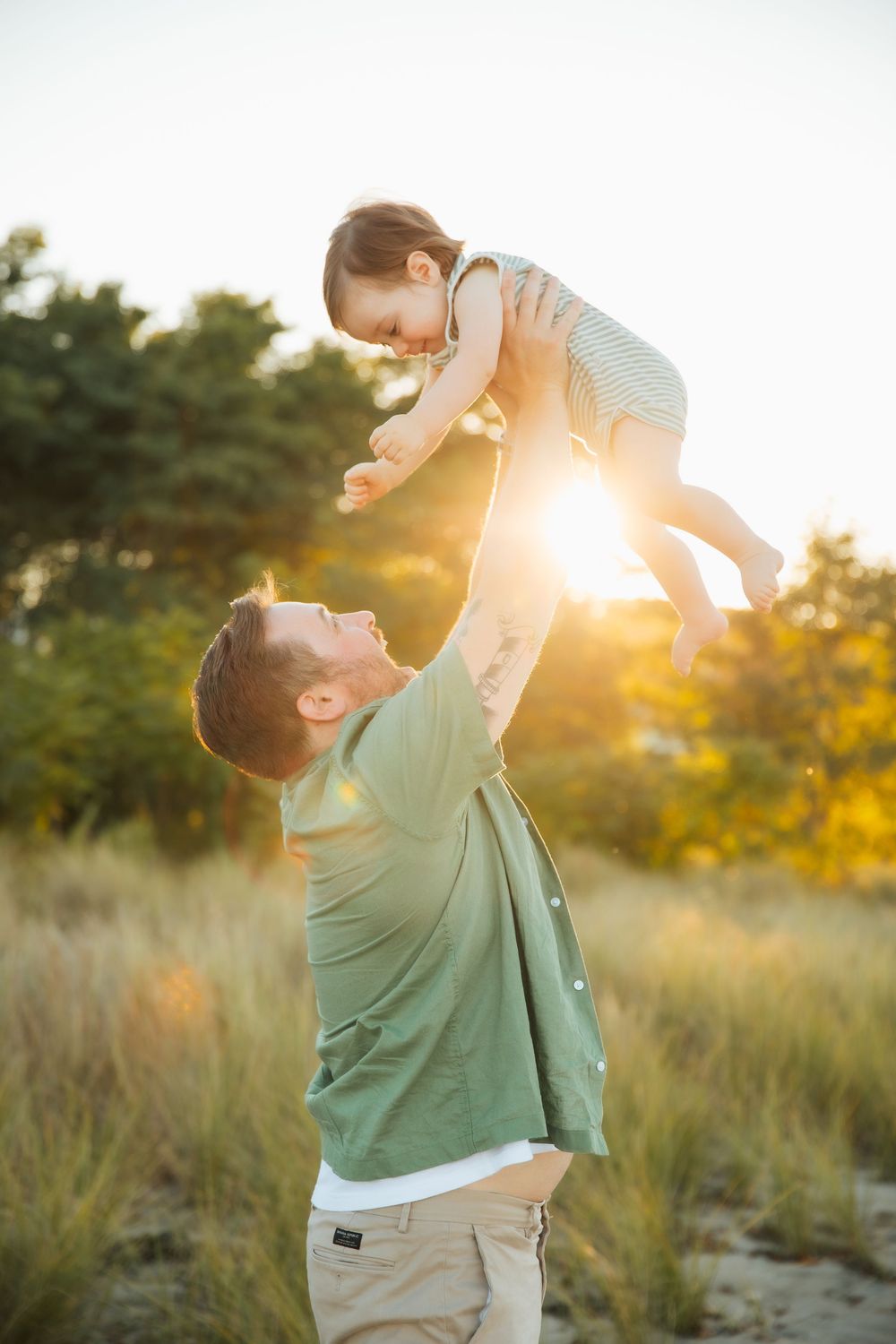 Father joyfully lifting baby in the air outdoors at golden sunset, surrounded by lush green trees and grass. Portsmouth, RI Rhode Island