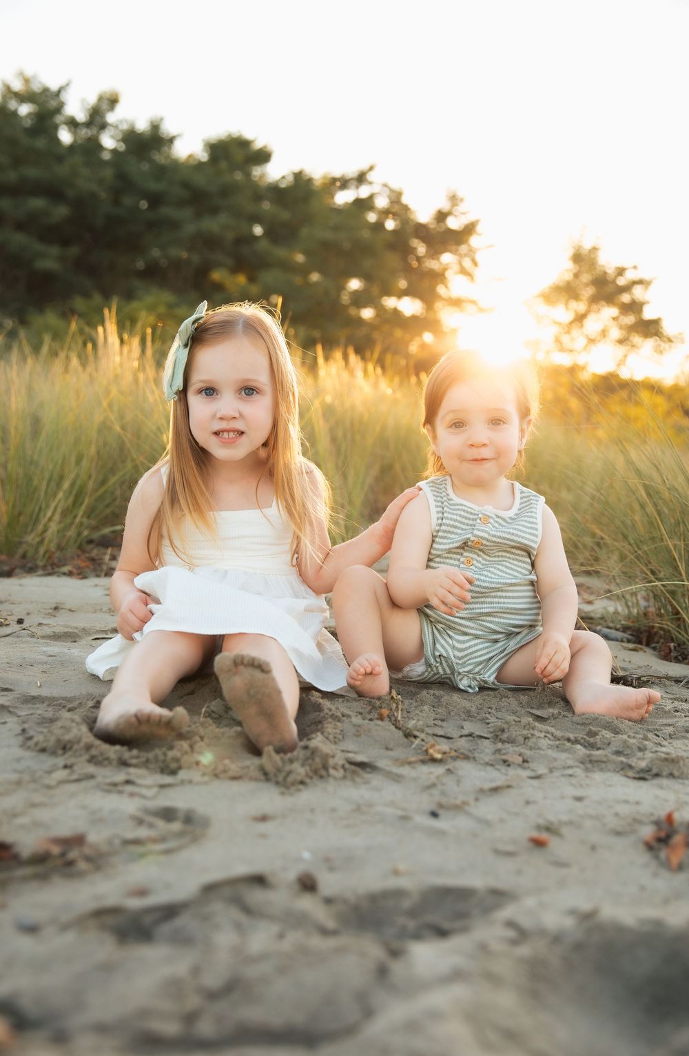 Two young children sitting on sandy beach at golden sunset, girl in white dress and toddler in striped romper. Portsmouth, RI Rhode Island