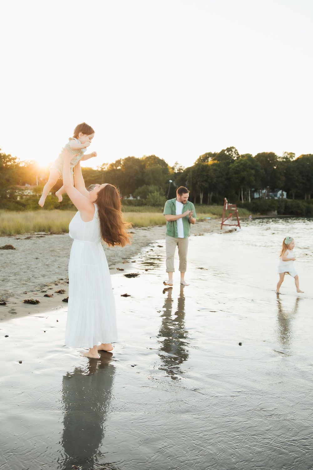 Family enjoying golden hour at the beach, mother lifting baby while father and child play in shallow water. Portsmouth, RI Rhode Island
