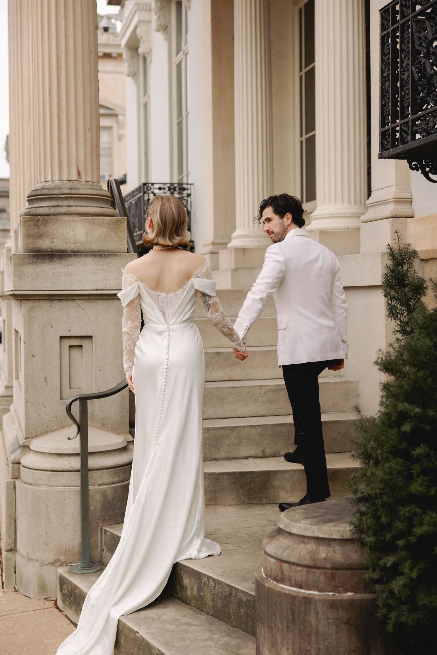 Bride in satin gown holds grooms hand ascending stone steps of elegant columned building.