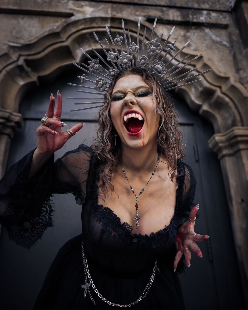 Female model in a dark blue dress and silver headpiece, portraying a vampire character with fangs and subtle blood, blending horror and beau