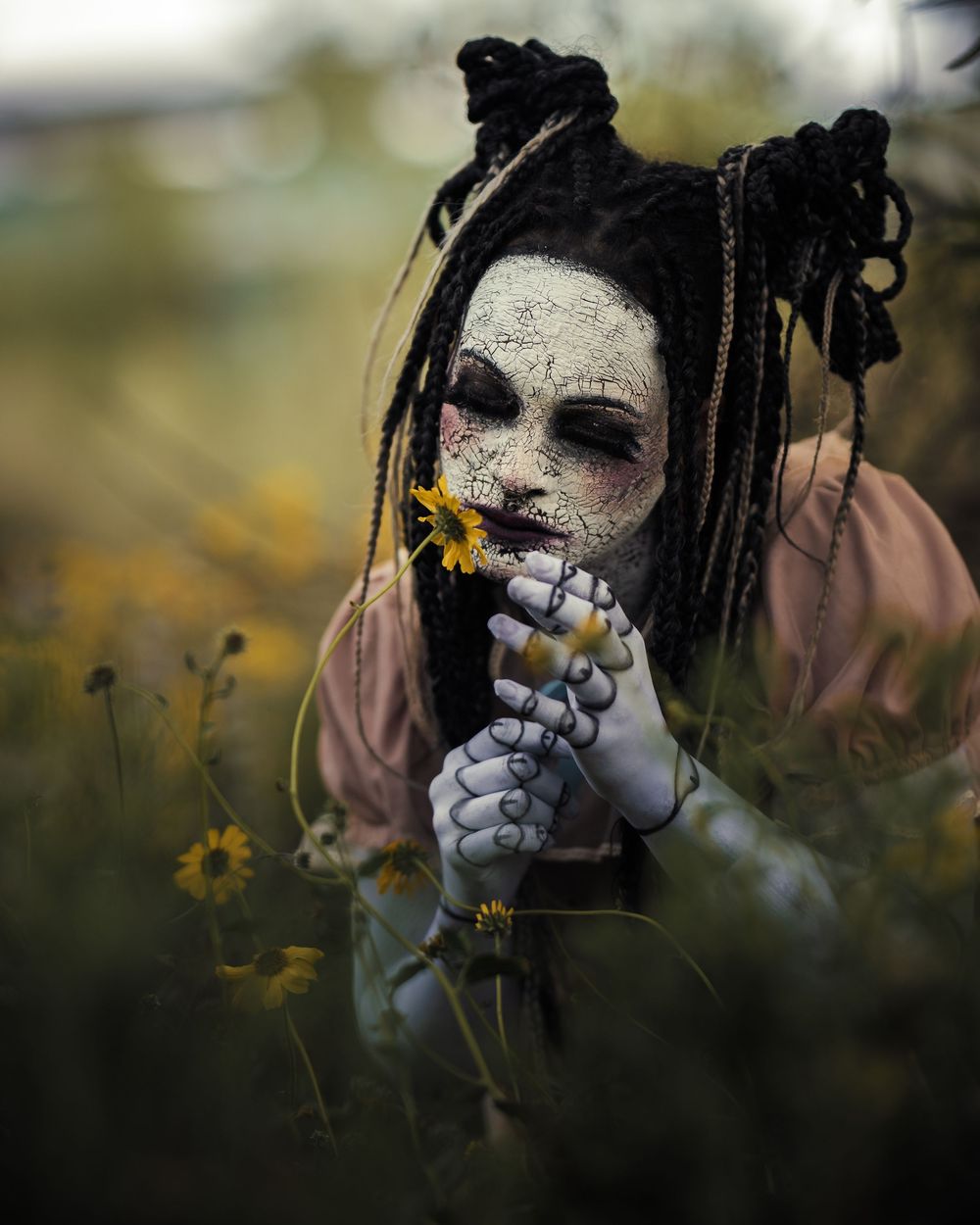 Model dressed as a cracked porcelain doll stares blankly while seated in a field, framed by dry grass and eerie natural light.