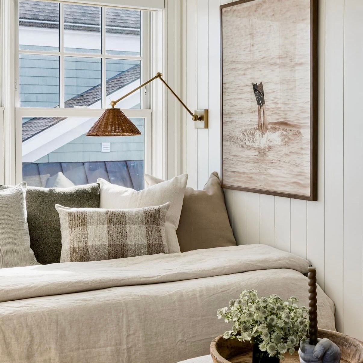 Cozy reading nook with linen daybed, plaid pillows, brass swing-arm lamp, and coastal wall art near a white window.