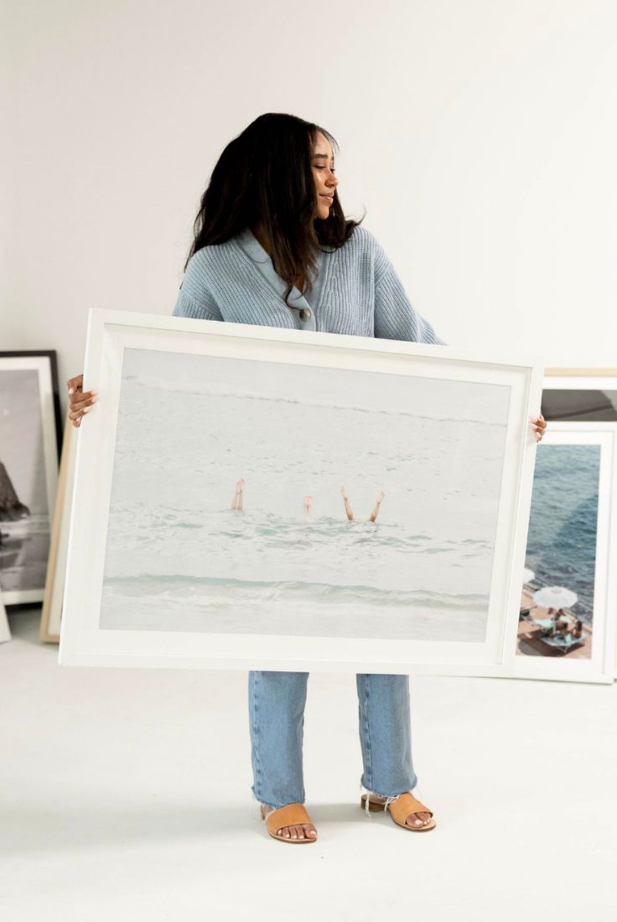 Someone in blue jeans and a sweater holds up a large framed beach photograph showing people swimming in the ocean.