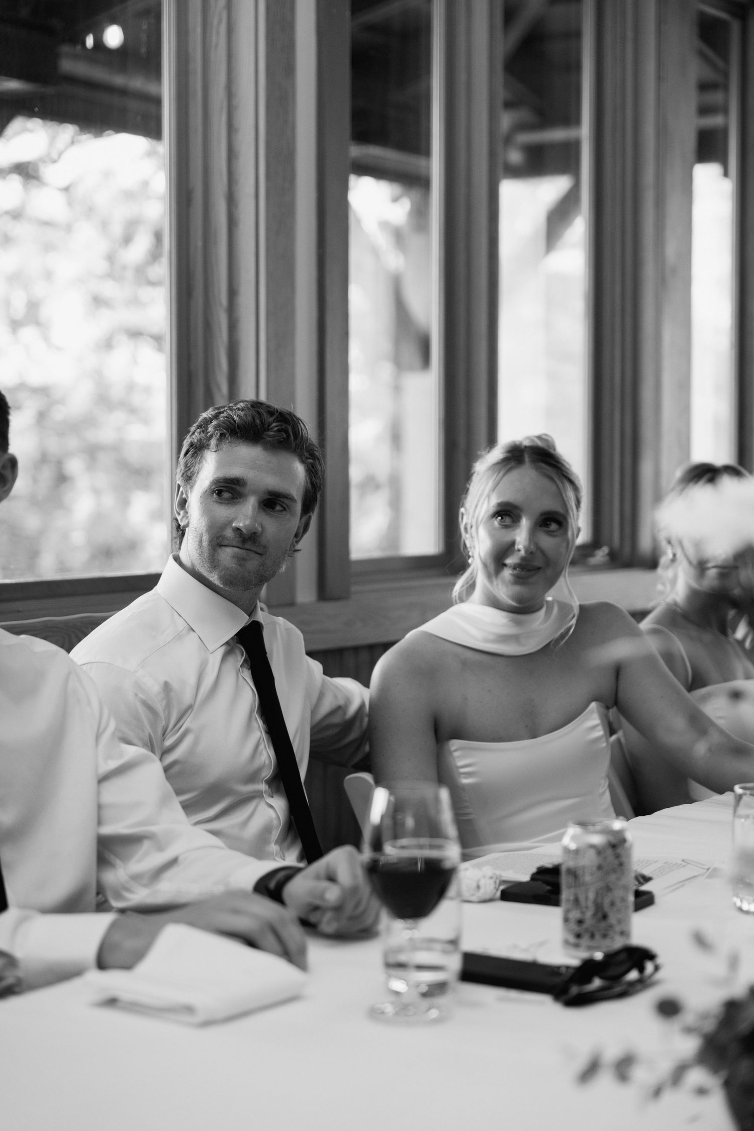 A black and white photo of guests at a wedding reception seated at a table with wine glasses.