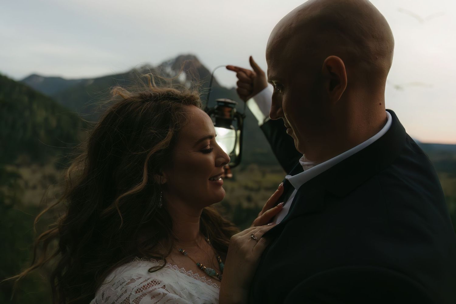 A romantic moment between newlyweds with windswept hair against a mountainous backdrop at sunset.