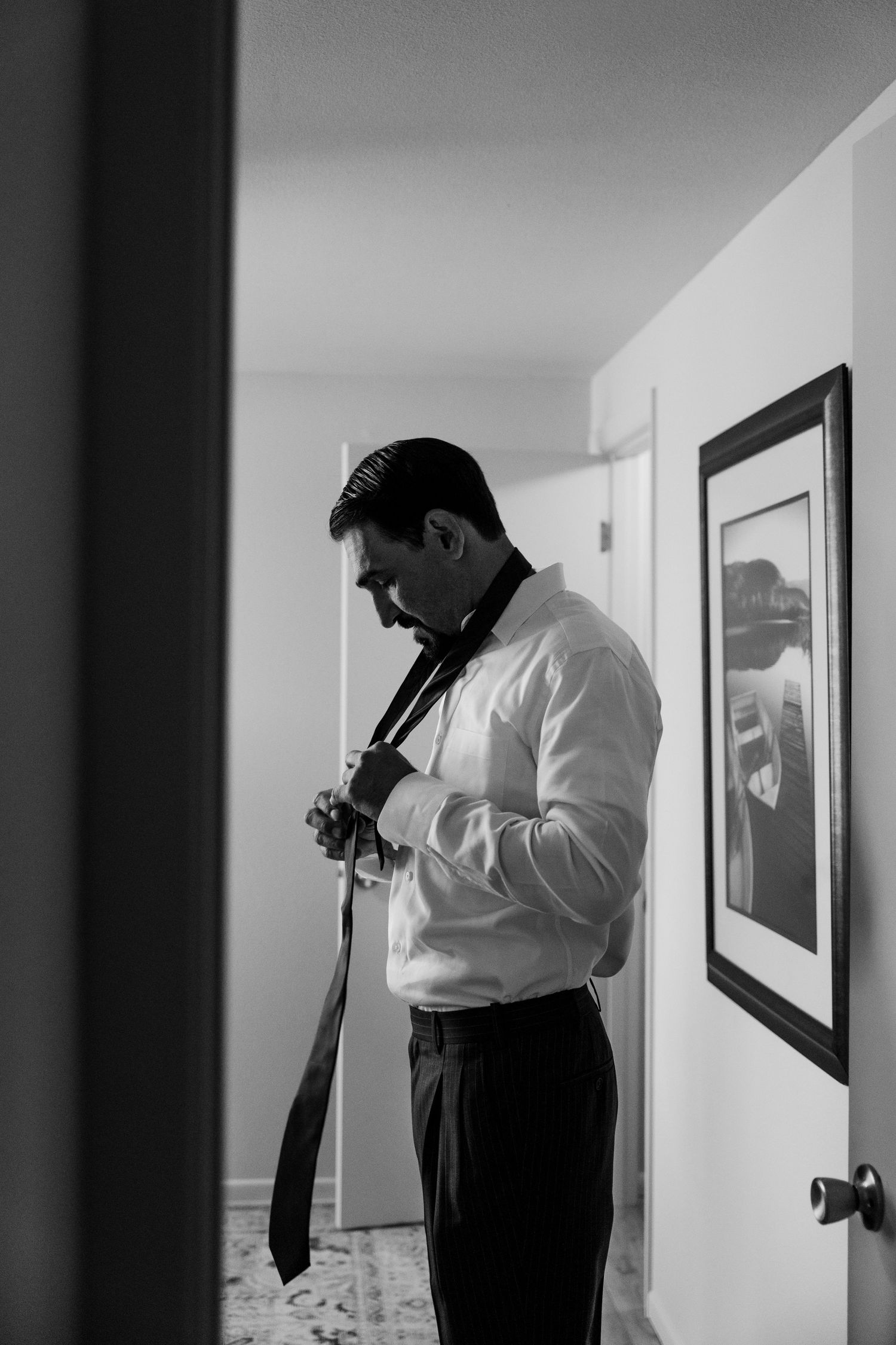 A man in a white dress shirt adjusts his tie while standing in a hallway with framed artwork on the wall.