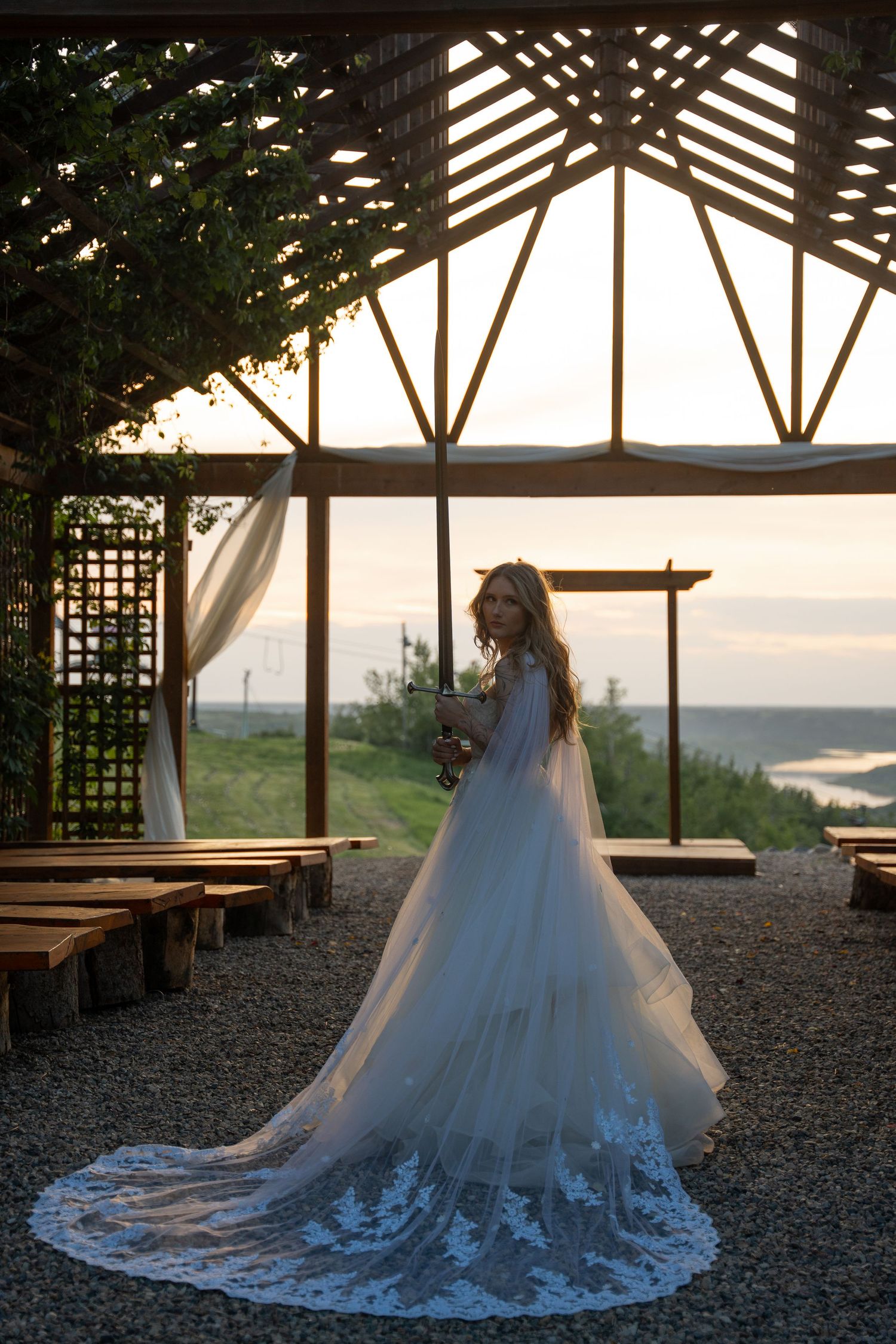 A bride in a stunning lace gown with long train poses in a rustic wooden pavilion at sunset.