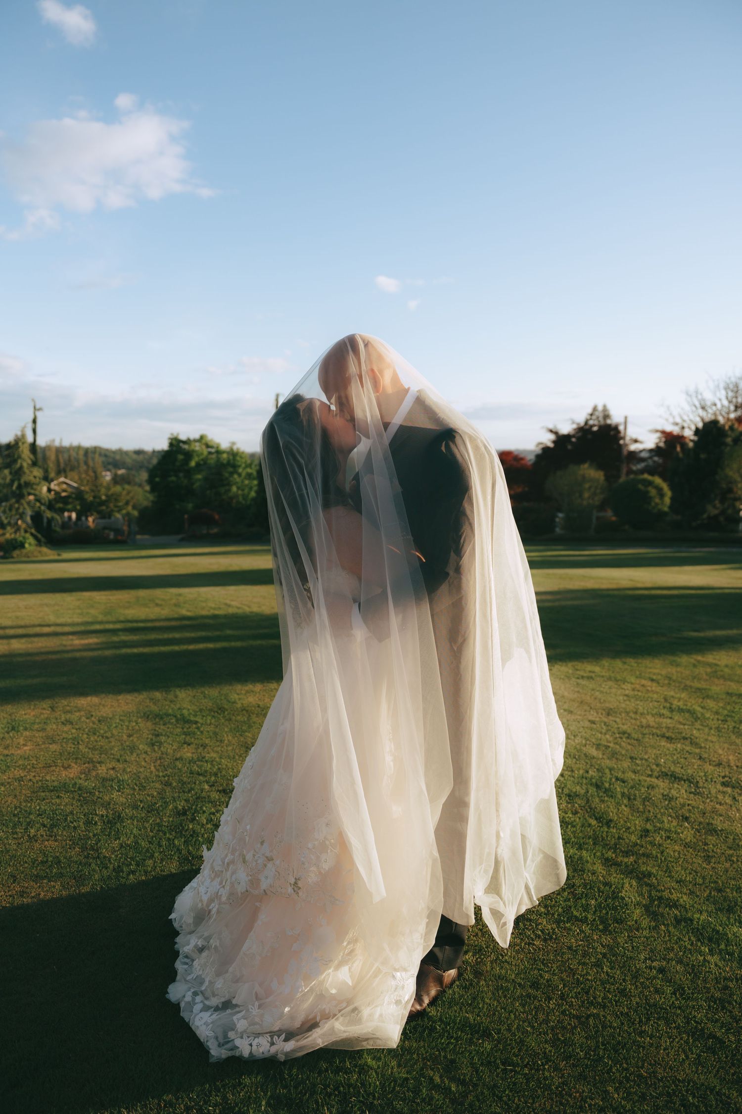 A couple in wedding attire share a romantic kiss under a flowing white veil while standing in a grassy field at sunset.
