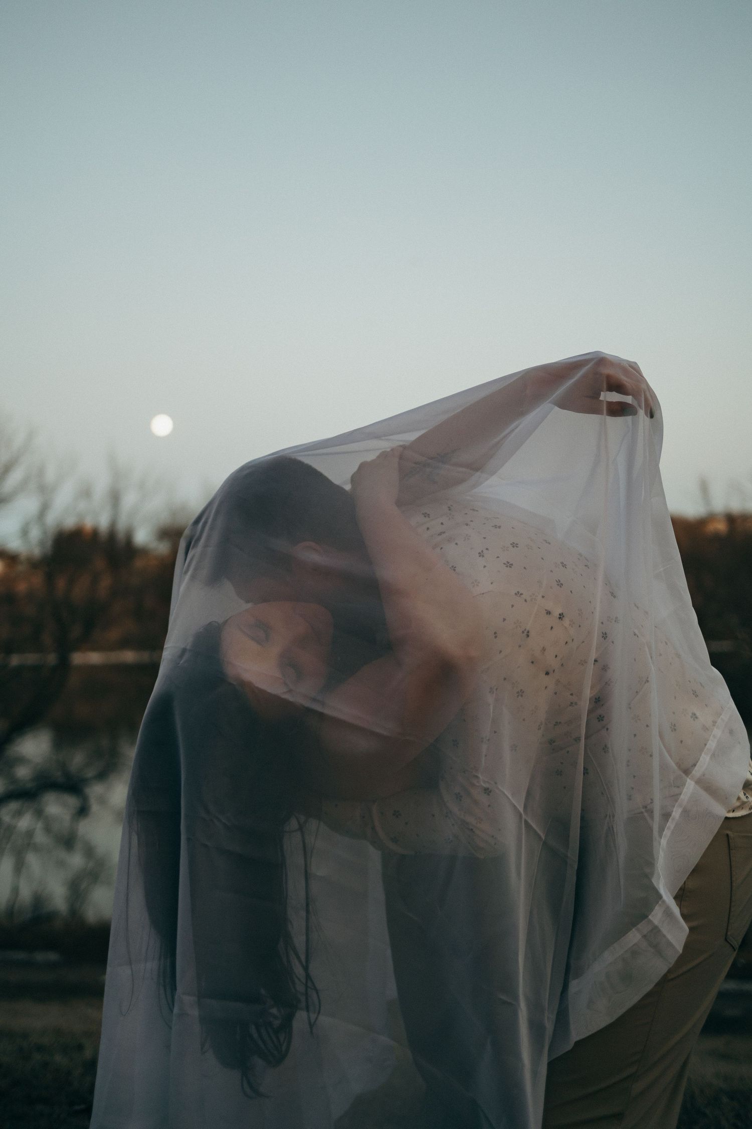 Ethereal figure draped in translucent veil stands against dusky sky with glowing moon in background.