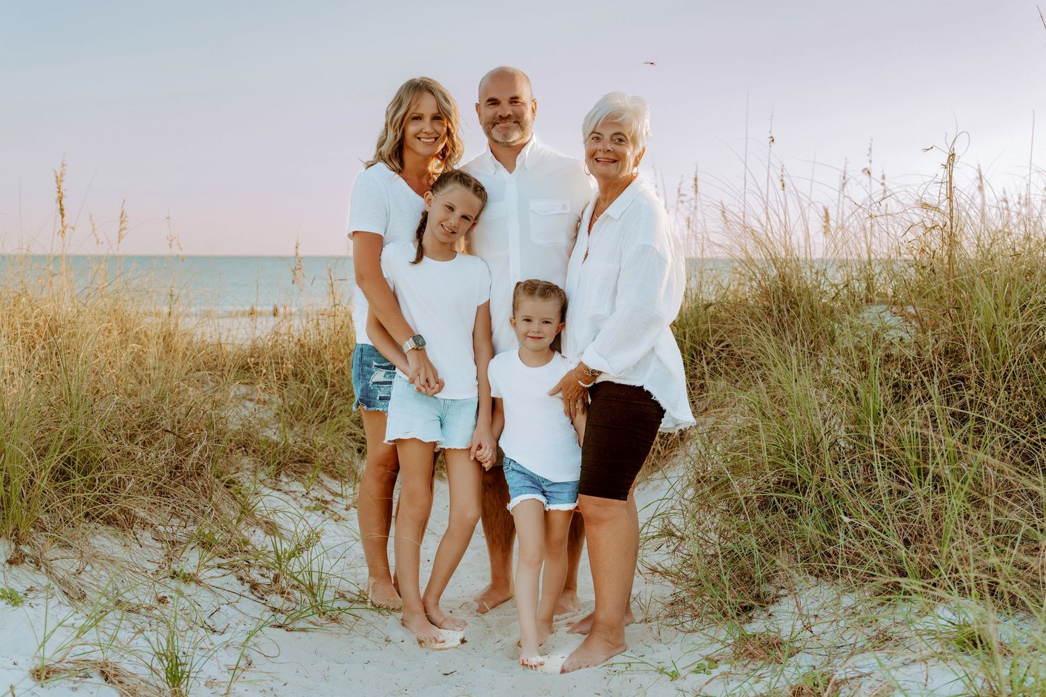 A family group poses together on a sandy beach path with ocean and dunes in the background at sunset.