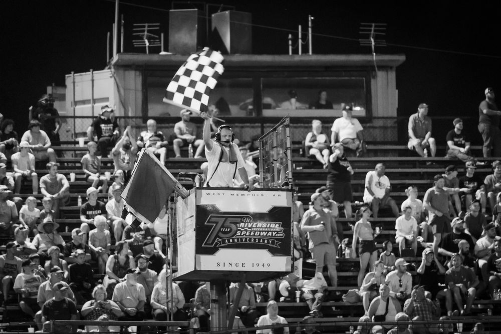 Black and white photo of racing fans in crowded stadium grandstands with checkered racing flags on display.
