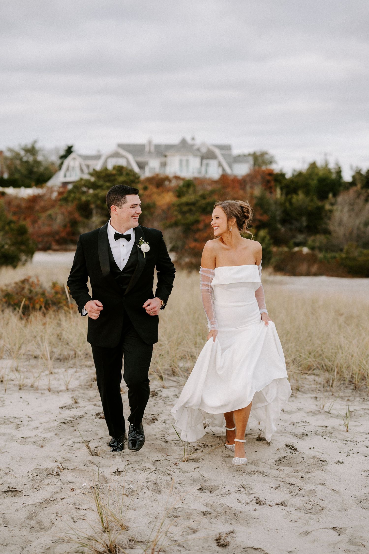 A couple in wedding attire runs playfully through sandy dunes near coastal homes.