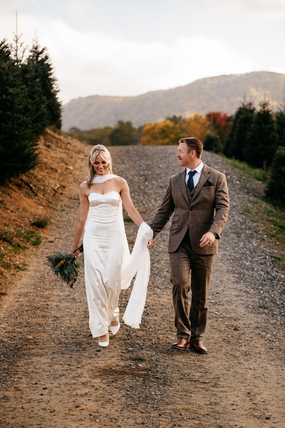 Bride and groom walk hand-in-hand on a gravel path surrounded by evergreens and fall foliage with mountains behind.
