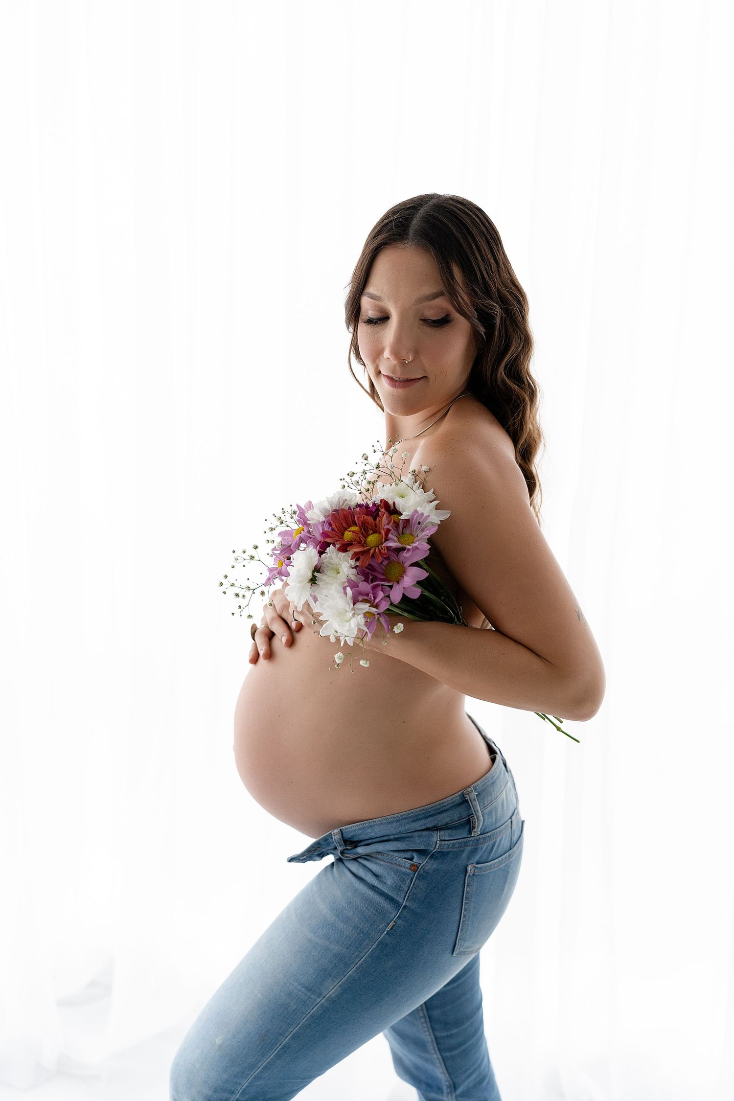 Expectant mother in jeans holds colorful flowers against her bare belly in a maternity photoshoot.