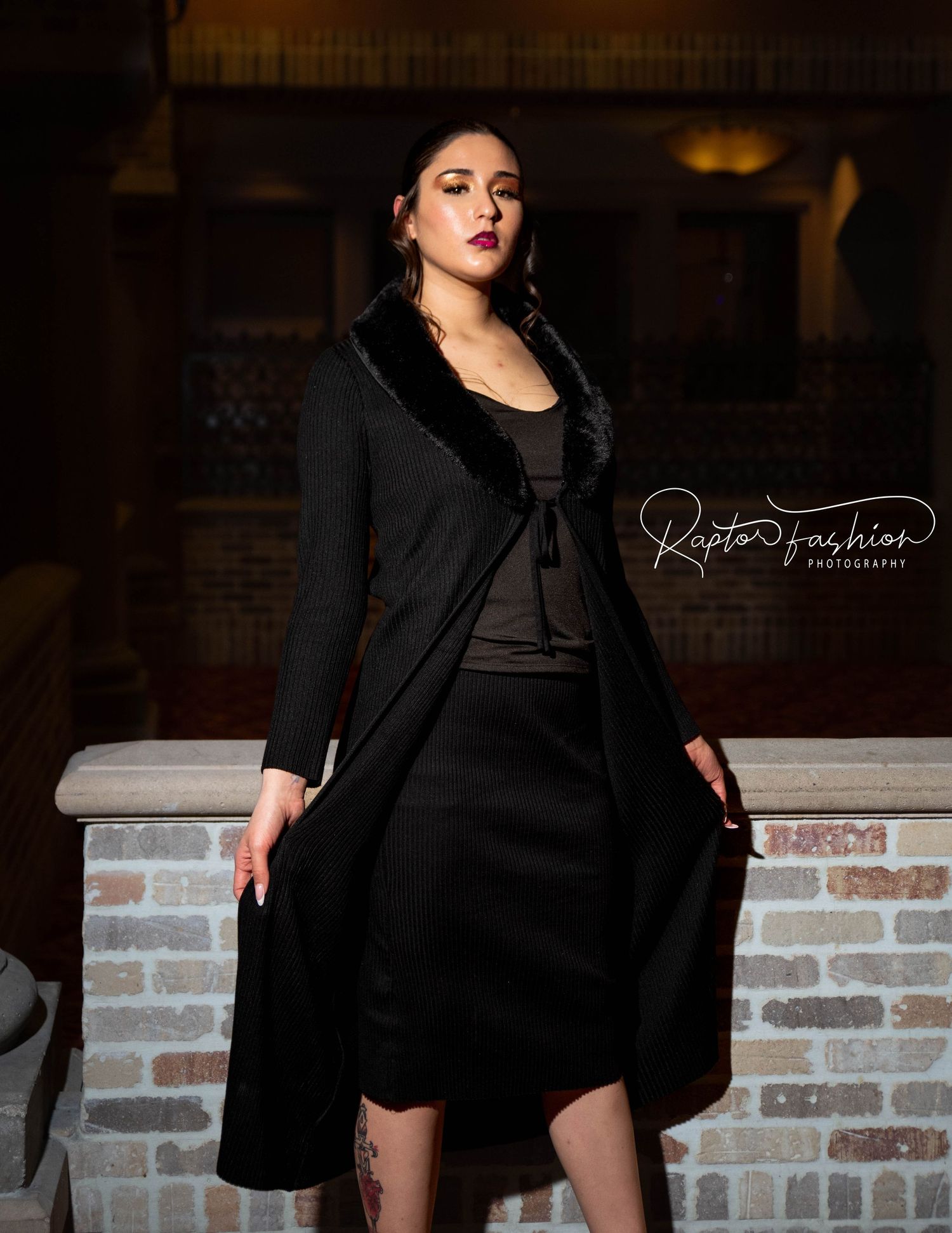 Fashion model in elegant black coat dress poses dramatically against brick wall in moody lighting.