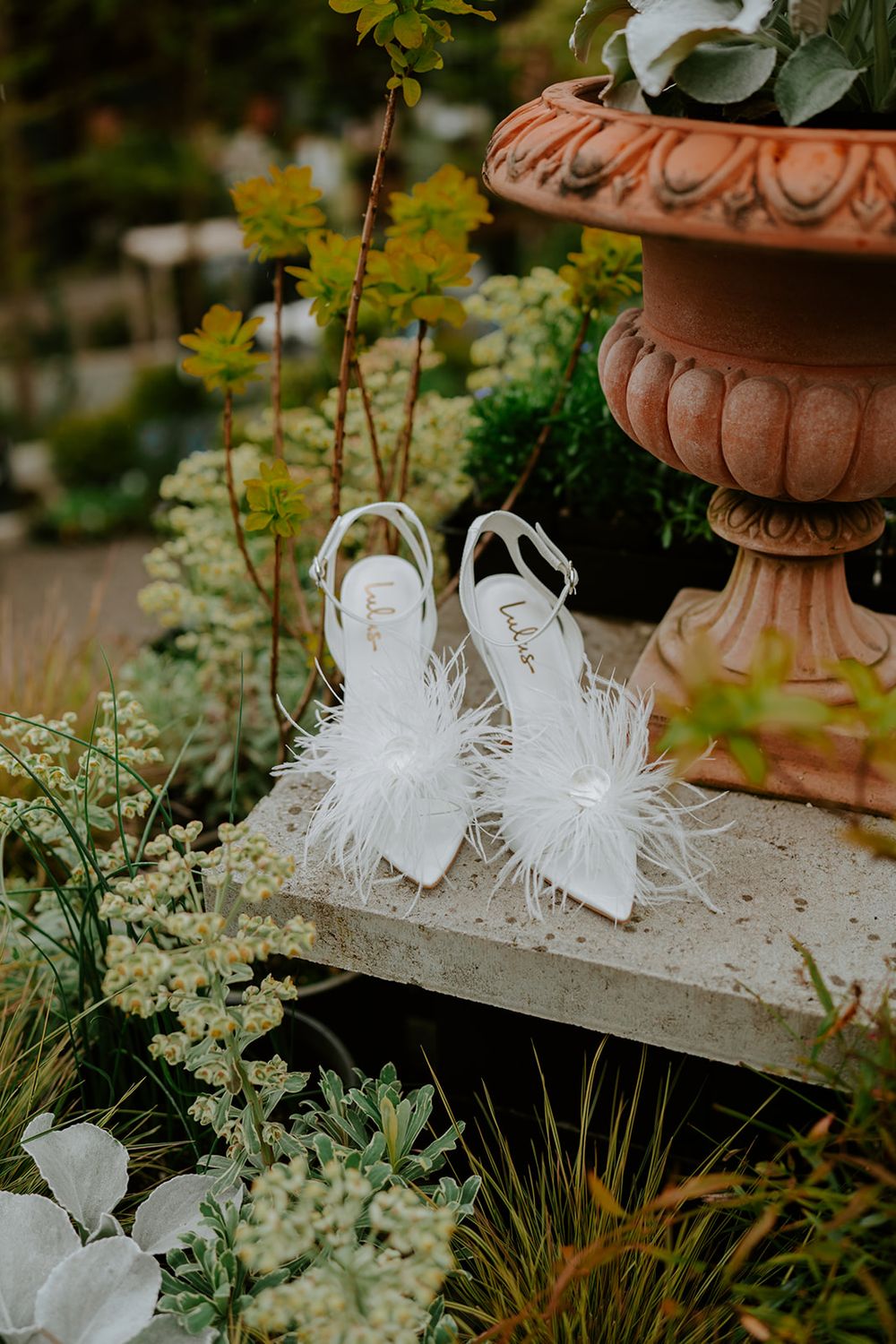 White feathered bridal sandals displayed near a terracotta garden urn amid lush greenery.
