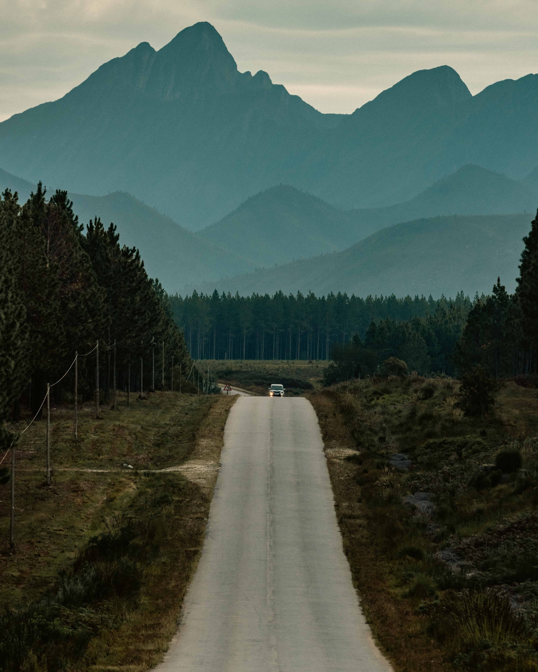 Long empty road leading towards misty mountain peaks with forest in the foreground.
