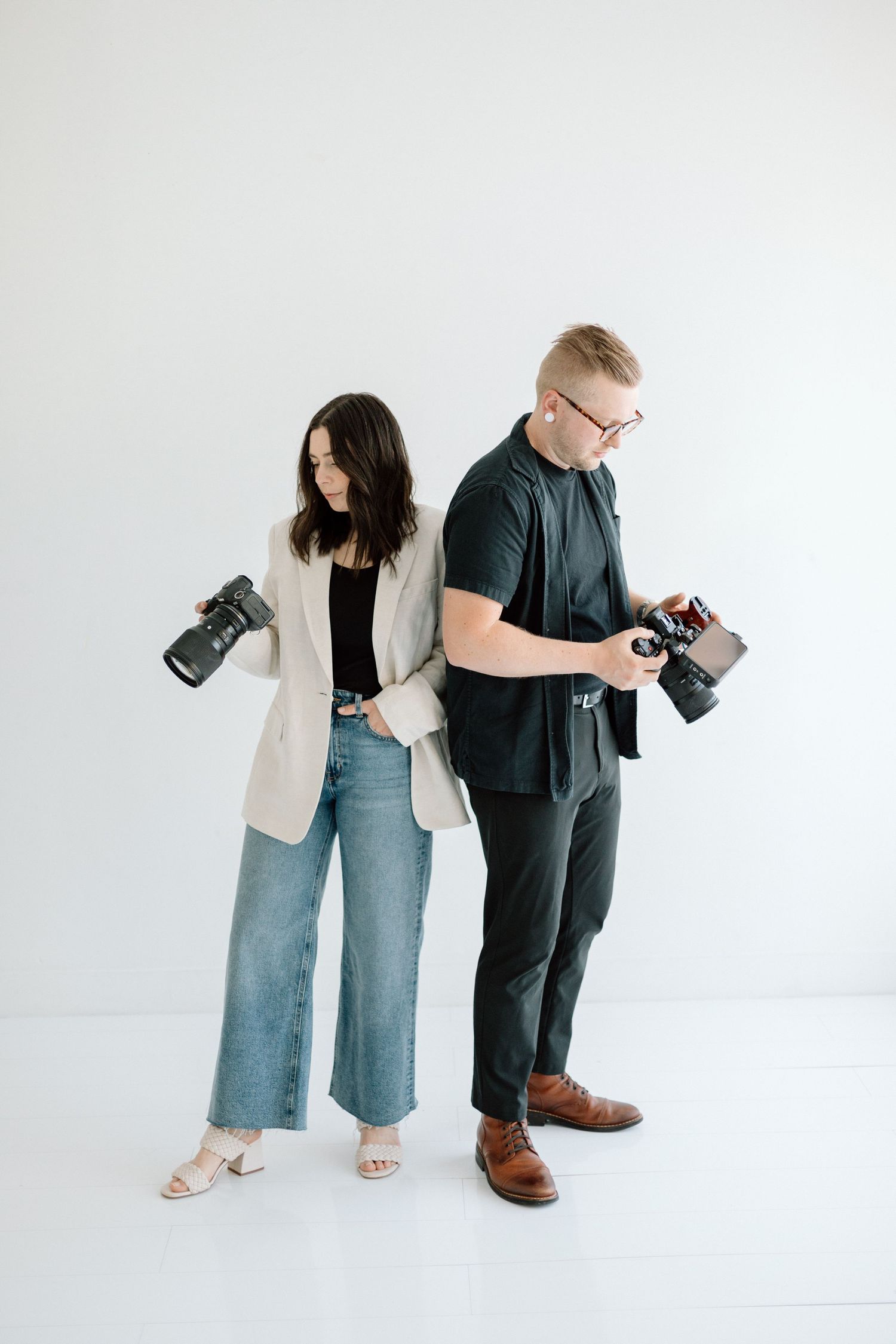 Two photographers holding cameras pose back-to-back against white background.