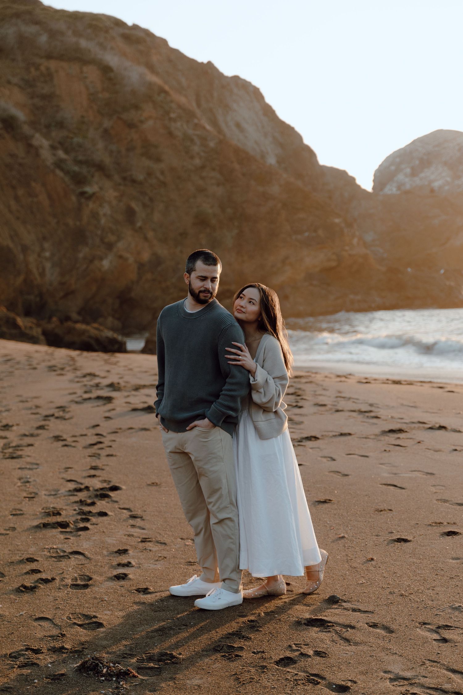 A couple embraces on a sandy beach with dramatic rocky cliffs in the background at sunset.