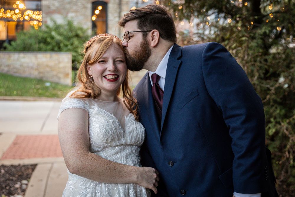 A joyful couple shares a sweet moment outdoors during their wedding, with the groom in navy suit kissing the bride in white lace.