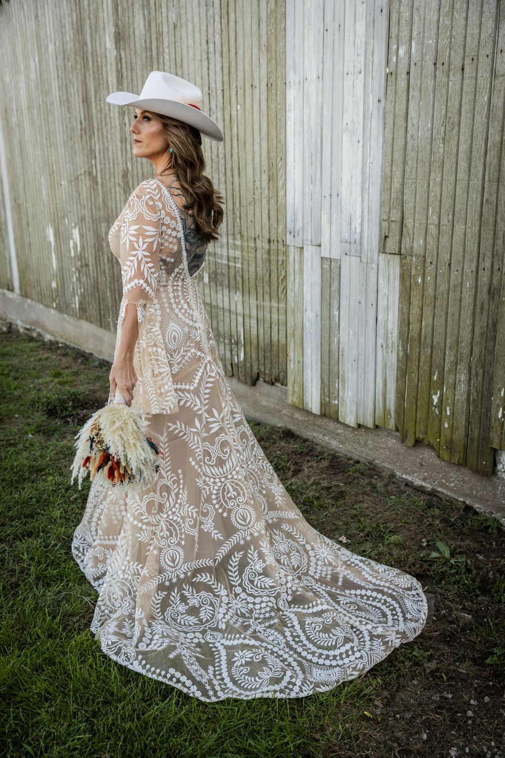 Person wearing an intricate lace wedding dress with flowing train against a wooden fence backdrop.