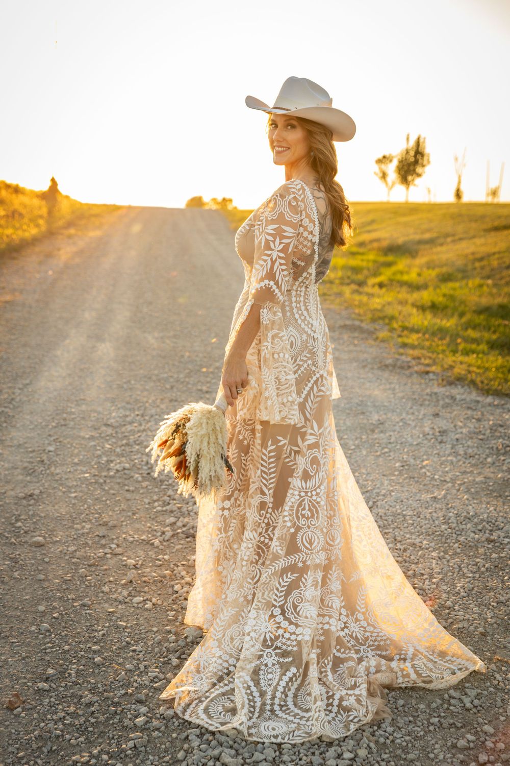 Bohemian bride in lace dress and cowboy hat twirls on dirt path at golden hour.