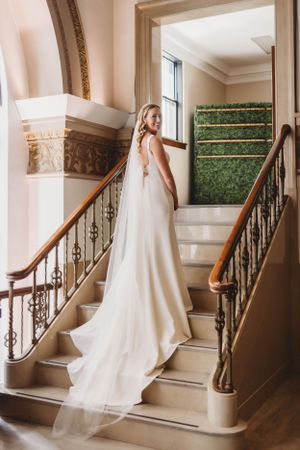 Bride in elegant white gown poses on grand staircase with ornate railing.