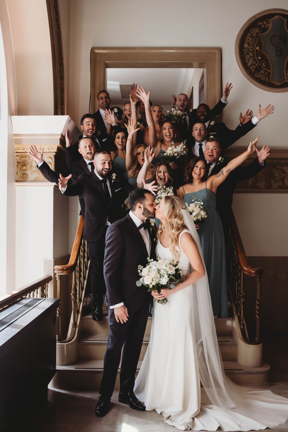 Wedding party celebrates joyfully on staircase behind newlywed couple.