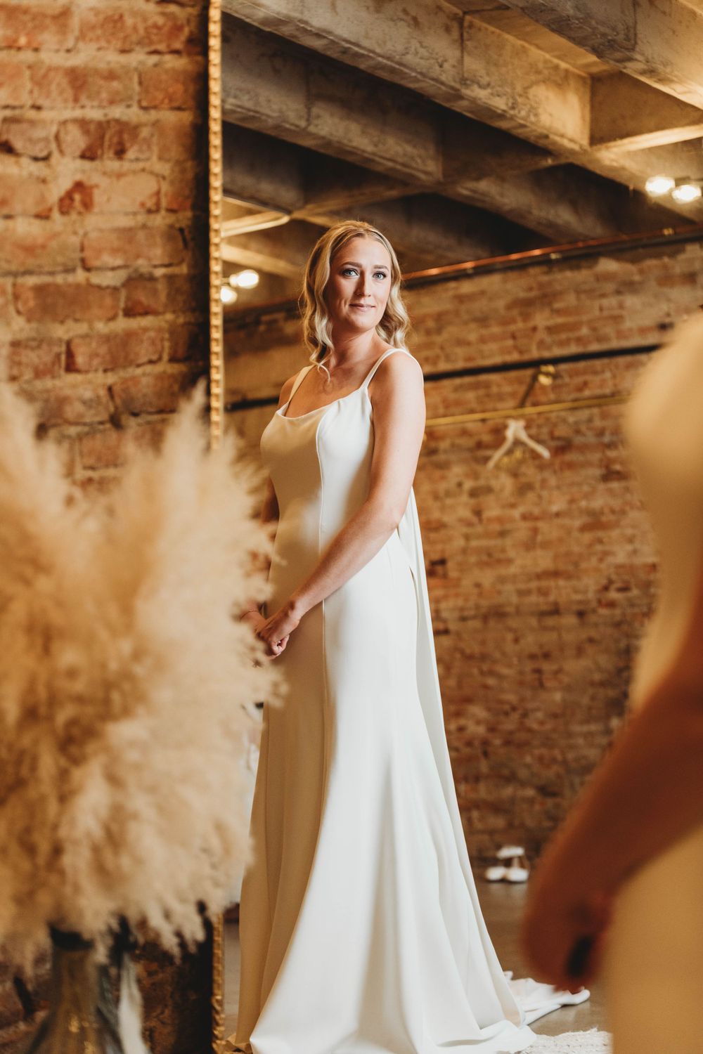 Bride in elegant white gown poses in industrial-style venue with exposed brick and pampas grass.