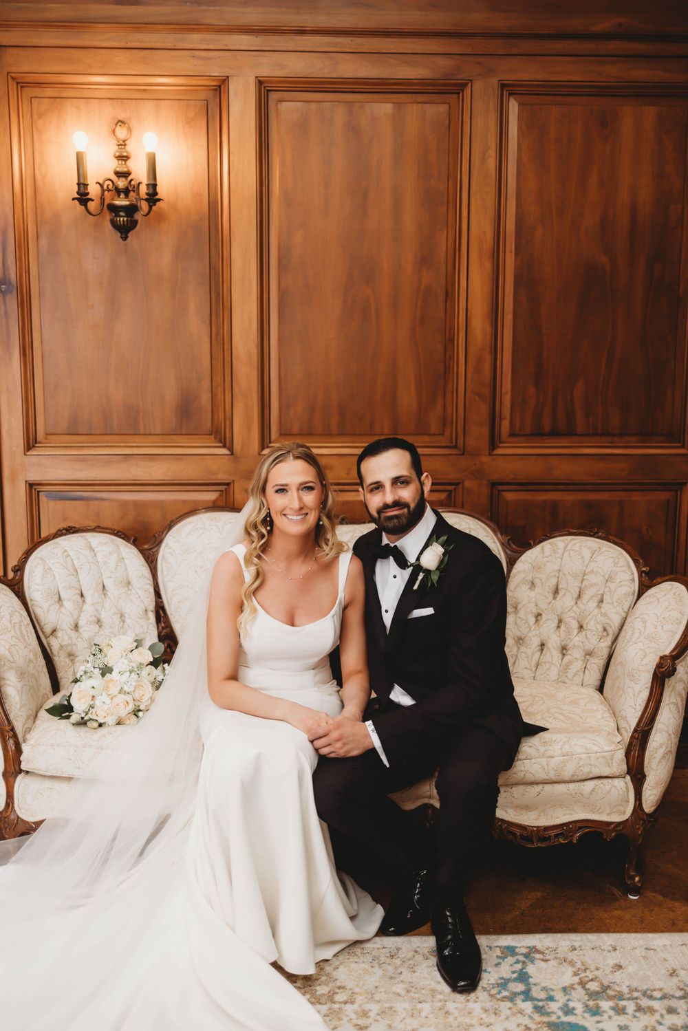 Newlyweds pose together on vintage tufted chairs against wood paneled wall.