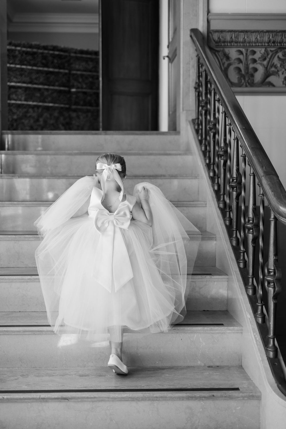 Young flower girl in white dress with large bow sitting on elegant staircase.