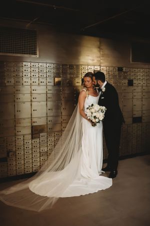Newlyweds pose intimately in front of vintage safety deposit boxes with bride holding white bouquet.