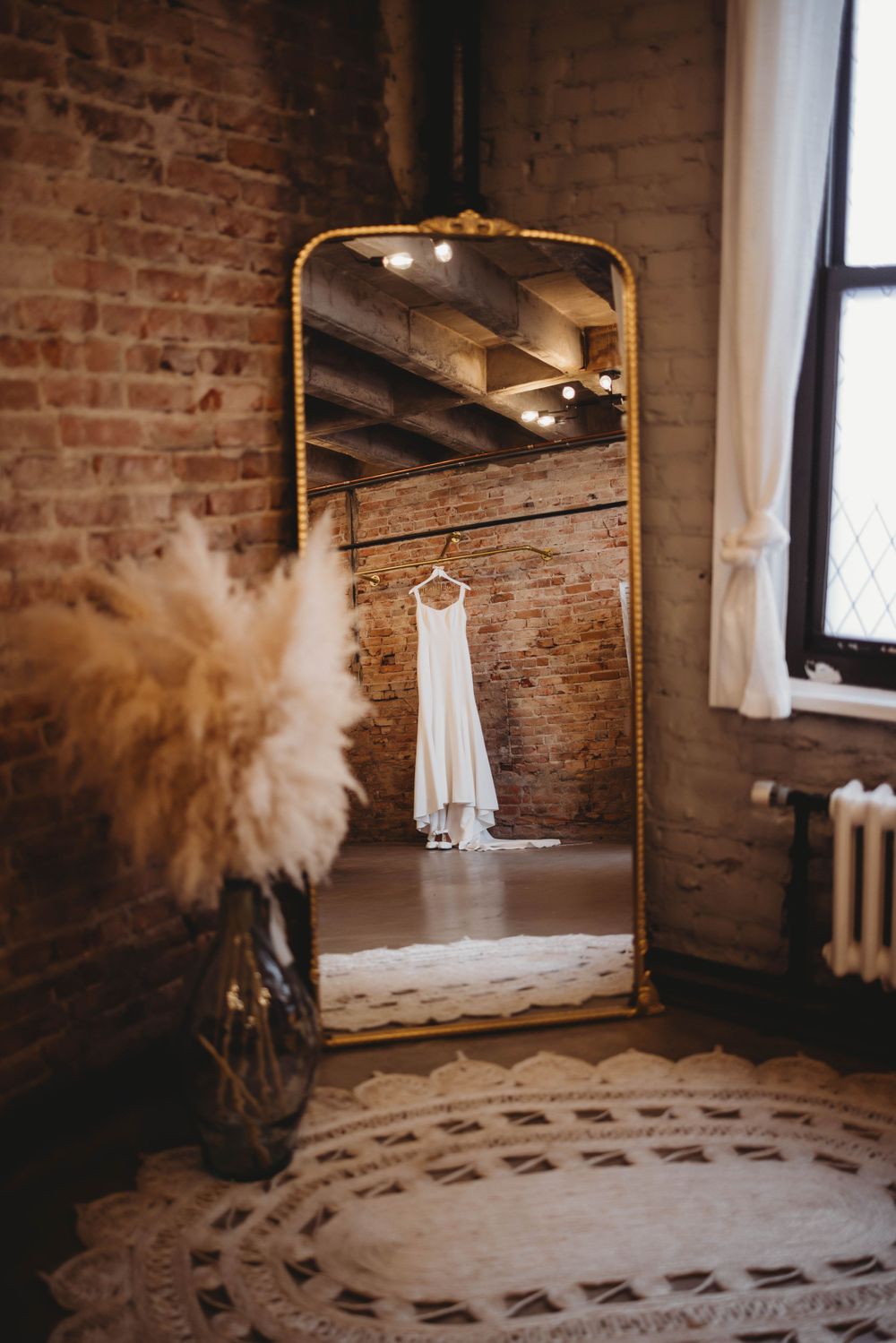 Gold framed mirror reflects a white wedding dress hanging against exposed brick walls with pampas grass decor.
